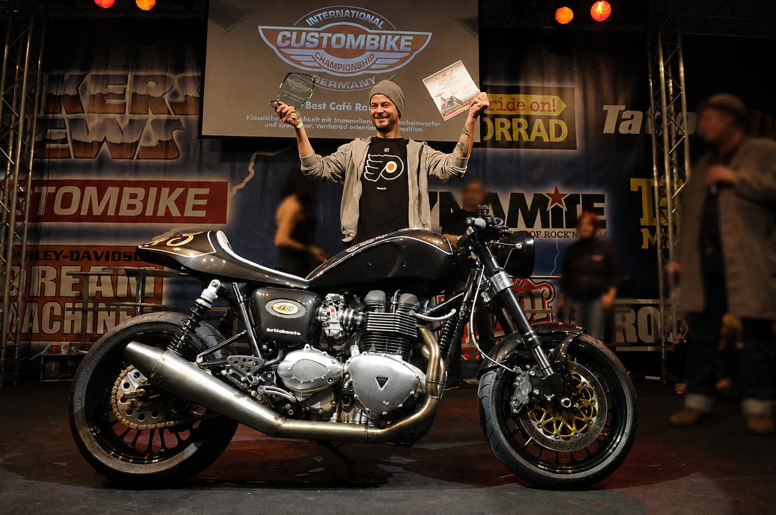 Arreno-Gründer Ivo Tschumi hebt Pokal und Urkunde über den Kopf, vor ihm auf der Bühne sein Café-Racer-Aufbau an der International Custombike Championship Germany.